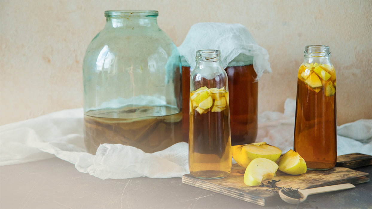 zwei große Einmachgläser eins mit Baumwolltuch zwei Flaschen mit Kombucha Apfelstücken auf Tisch